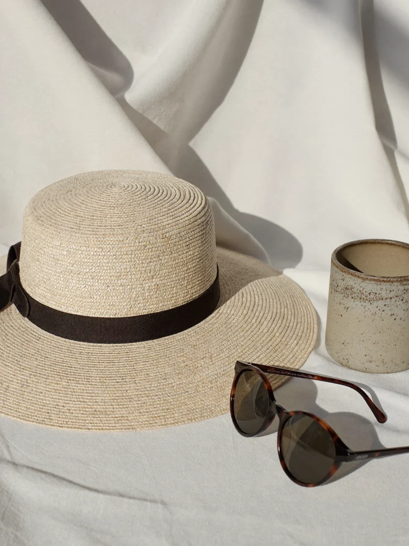 Strohhut mit dunklem Band auf Leinen neben Tasse und Sonnenbrille